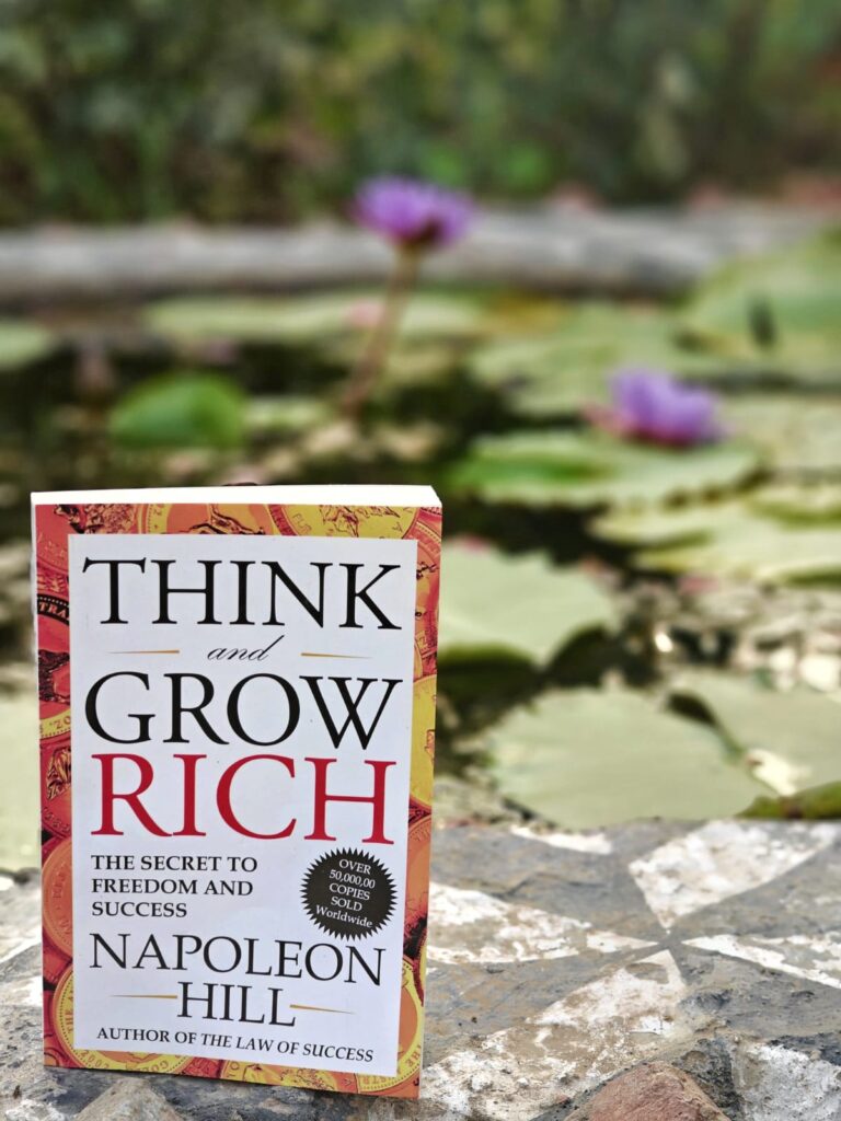 The book "Think and Grow Rich" near a pond with lotus in the background.